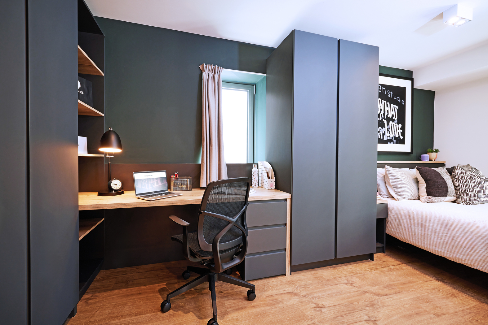 Modern bedroom with a workspace featuring a wooden desk, laptop, and black chair. Dark gray wardrobes and shelves flank the desk. A bed with patterned pillows is in the corner. The room has wood flooring and a window with curtains.