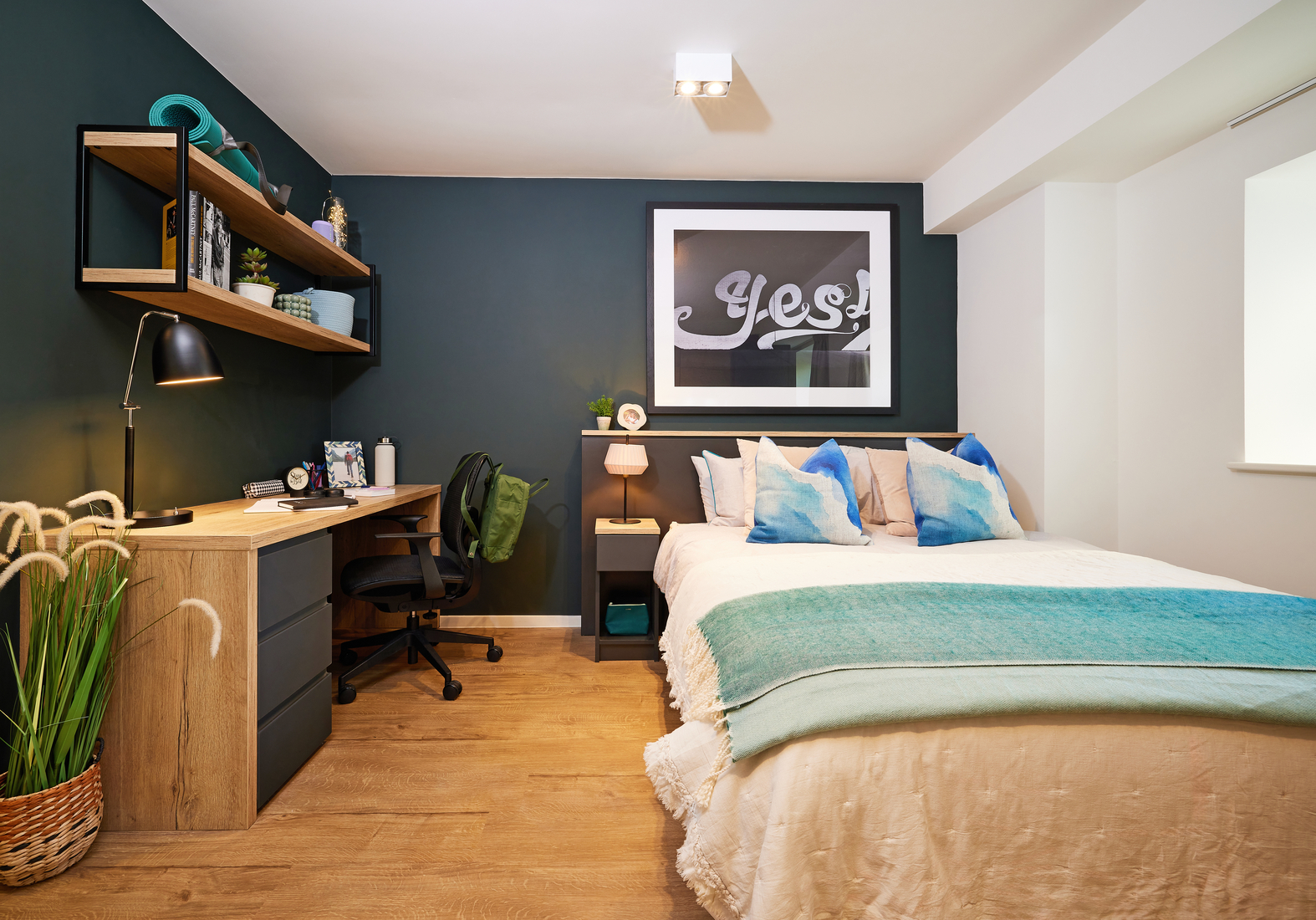A modern bedroom with a bed featuring white bedding and blue throw pillows. A dark accent wall displays a framed "Yes" artwork. There's a wooden desk with a black chair, shelves, a lamp, books, and a plant on a wooden floor.