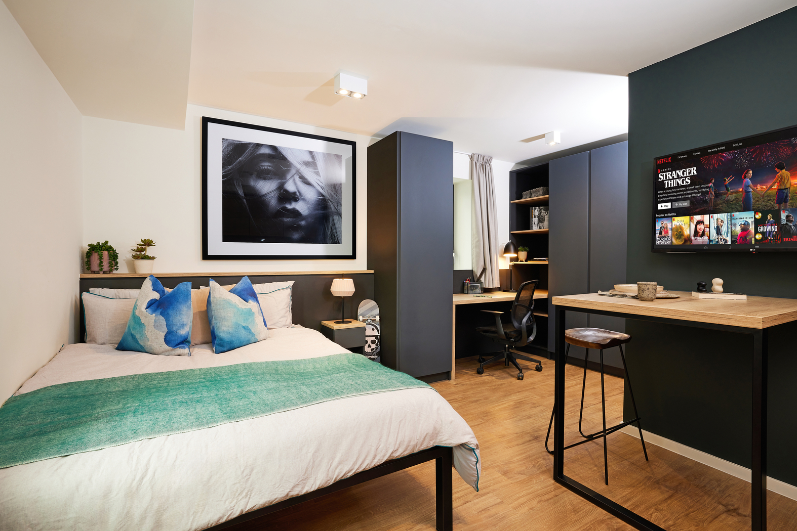A modern bedroom with a neatly made bed, blue and turquoise pillows, and a large framed portrait on the wall. A desk with a chair is in the corner, and a wall-mounted TV displays a streaming service interface.