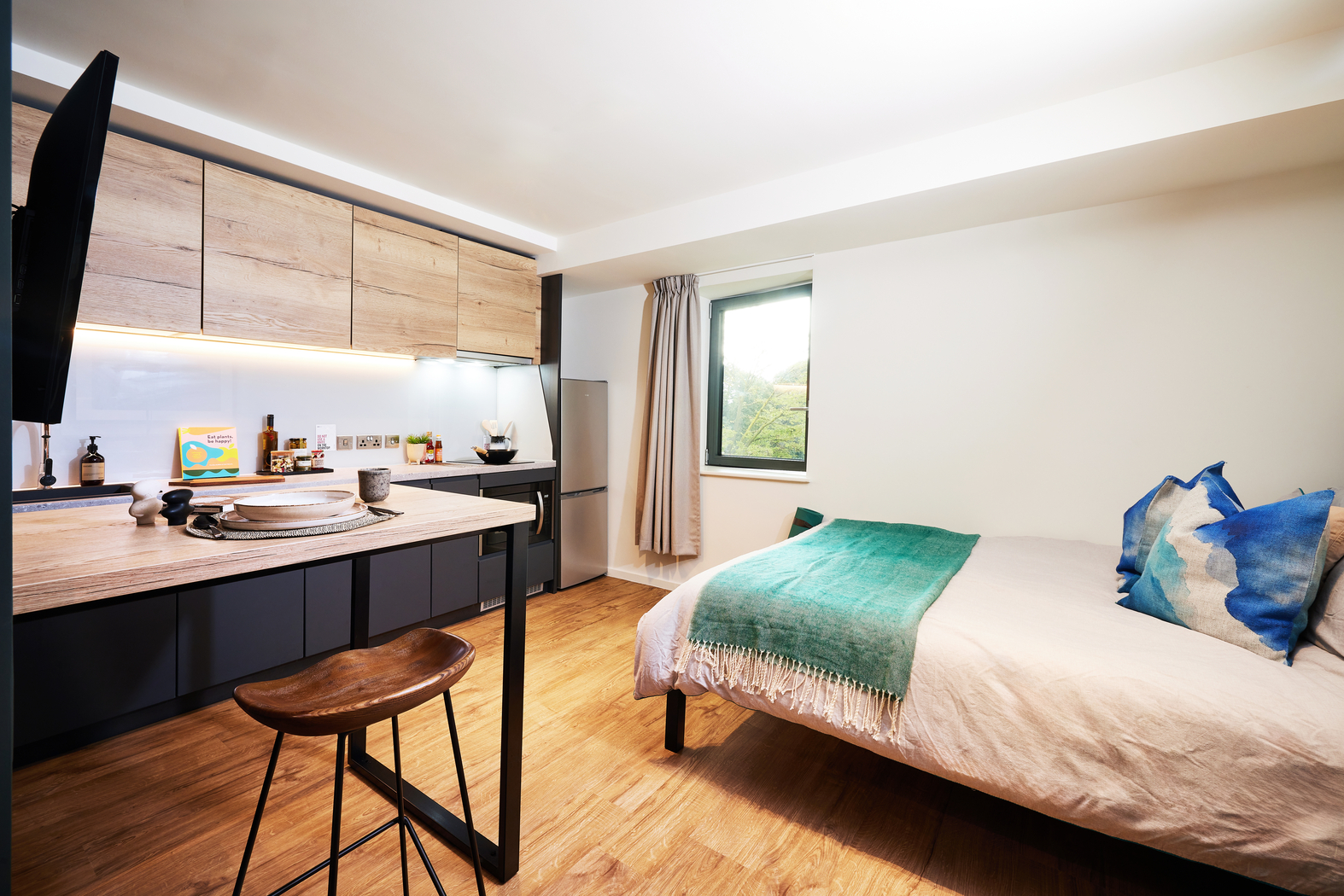 A modern studio apartment features a bed with blue and green bedding, a wooden bar stool, and a kitchenette with wooden cabinets. A window with curtains allows natural light, highlighting the wooden floor and minimalist decor.