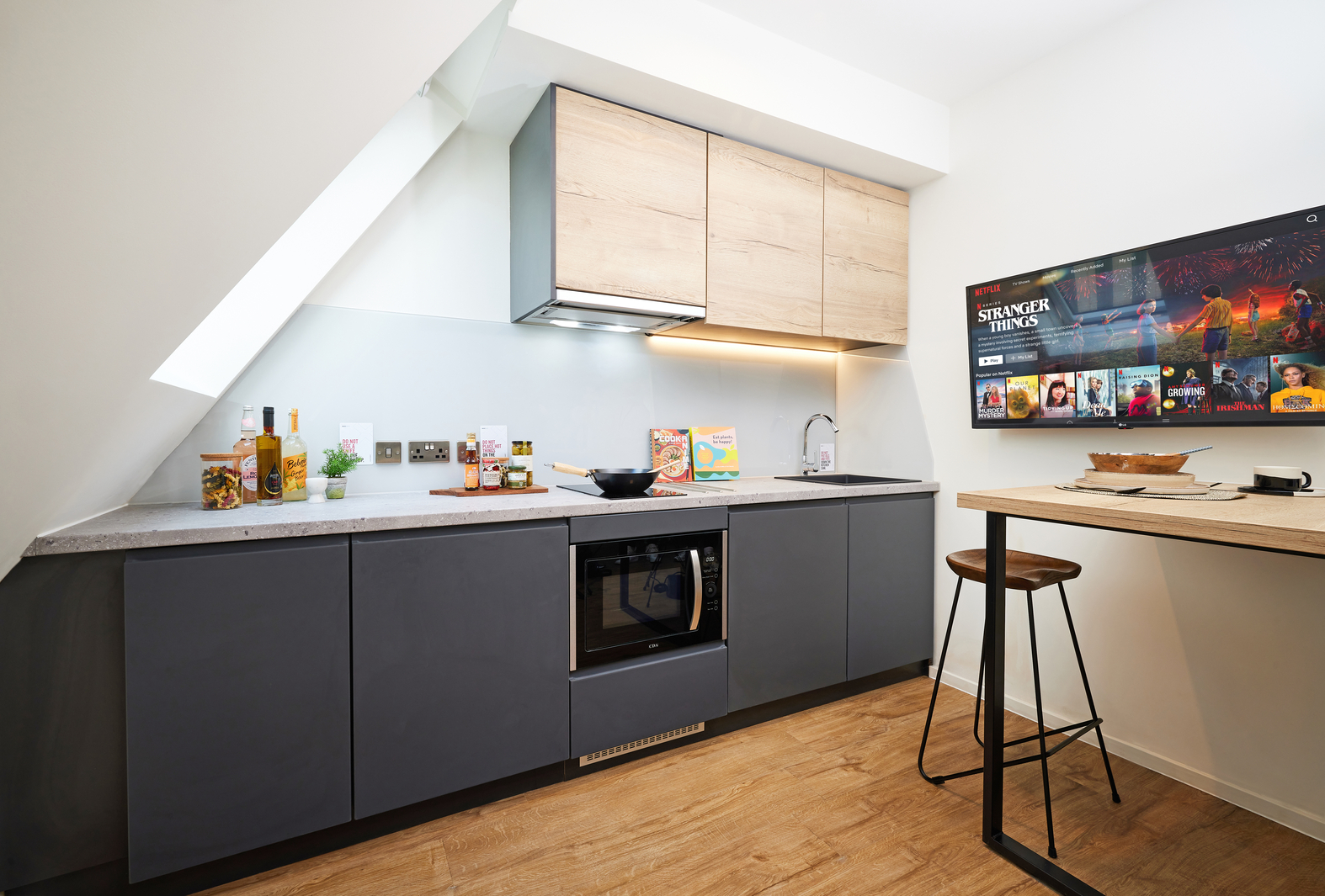 Modern kitchen with dark lower cabinets, light wood upper cabinets, and a marble countertop. A wall-mounted TV shows a streaming app. A wooden bar stool stands next to a tall table. Ambient lighting and a slanted ceiling add a cozy feel.
