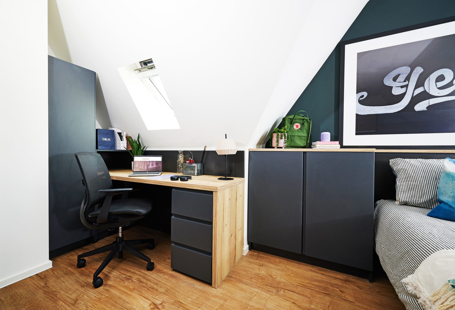 A cozy home office under a slanted ceiling, featuring a wooden desk with a dark chair, a laptop, and decorative items. There's a cabinet with storage boxes and a green bag. A large framed artwork hangs on the wall next to a small bed with cushions.