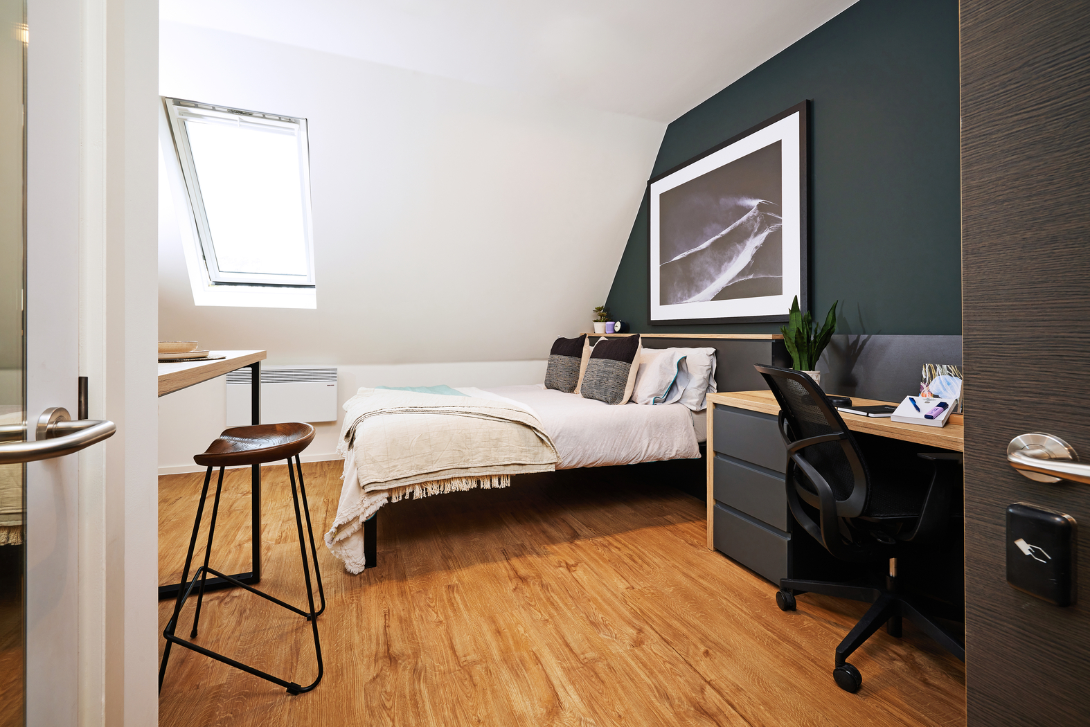 A modern bedroom with a sloped ceiling features a neatly made bed with cushions, a large framed art piece above, a wooden desk with a stool, a potted plant, and a small black office chair near a dresser. A skylight brightens the room.