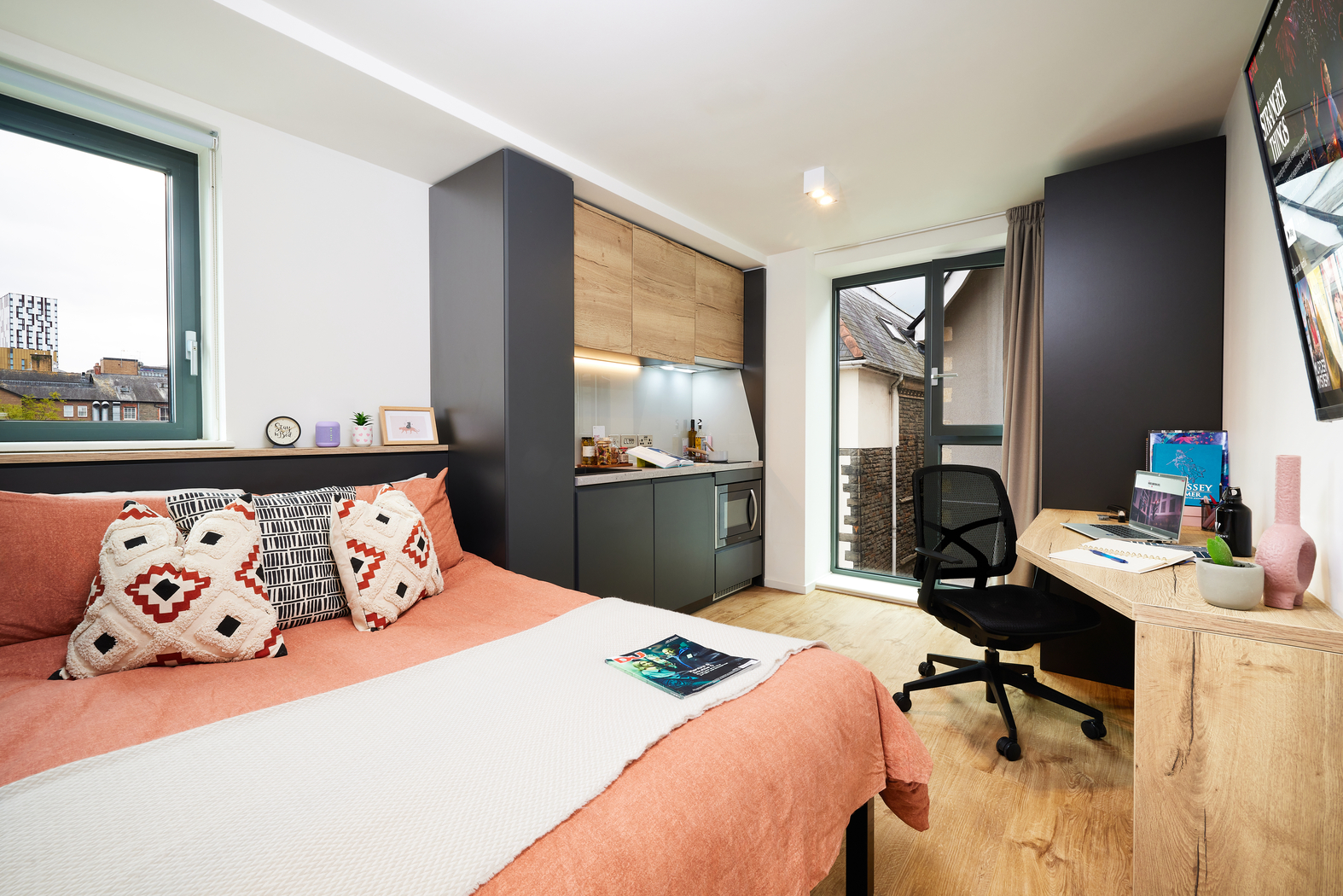 A modern studio apartment with a cozy bed featuring decorative pillows and a throw, a compact kitchenette, a work desk with a chair, and a large window next to a glass door leading to a balcony. Light wood flooring and subtle decor add warmth.