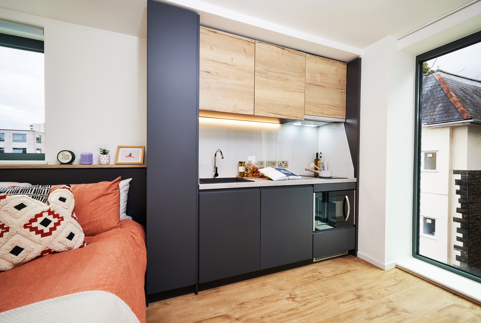 A modern studio apartment with a compact kitchen featuring dark cabinets, wood-accented upper cupboards, and a small counter. An orange sofa with a patterned cushion is on the left, and large windows on the right provide natural light and views outside.
