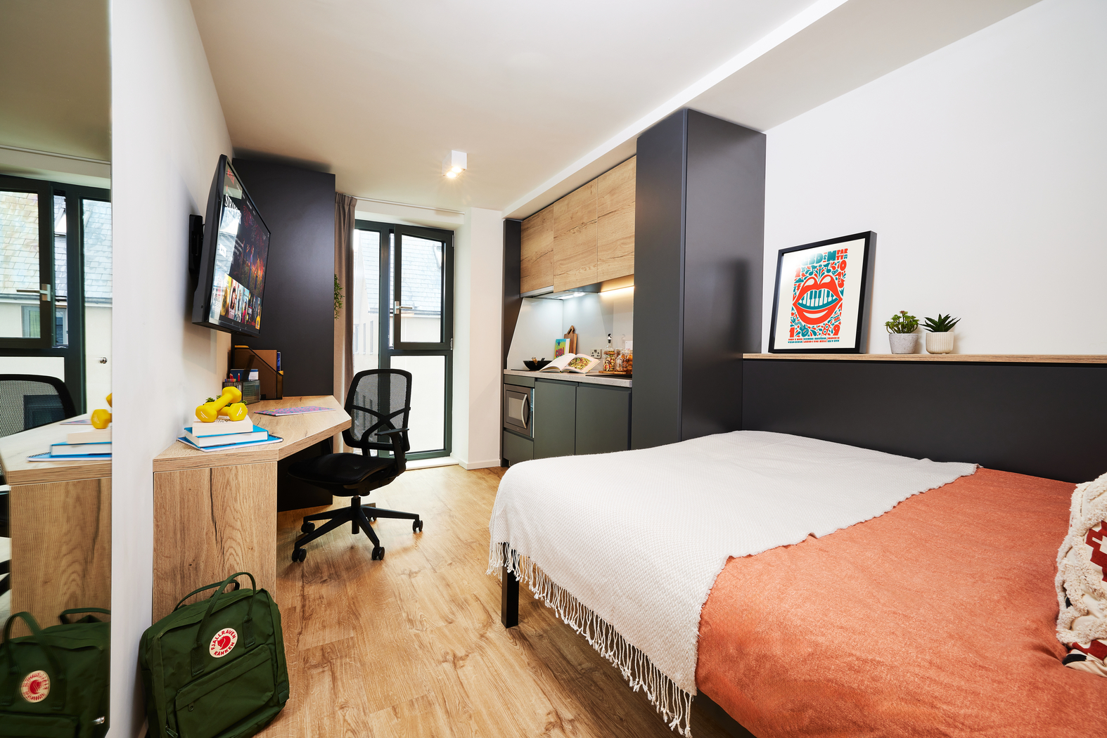 Modern dorm room with a bed, orange blanket, and pillows. A sleek desk and chair face a wall-mounted TV. Kitchenette with storage, plants, and artwork above. Large window with daylight. Two backpacks on the floor. Bright, organized space.