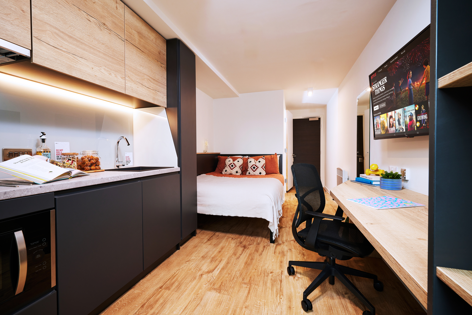 A compact studio apartment with wooden flooring features a kitchenette, a bed with heart-patterned pillows, a flat-screen TV, and a work desk with a black chair. The space is neatly organized and brightly lit.