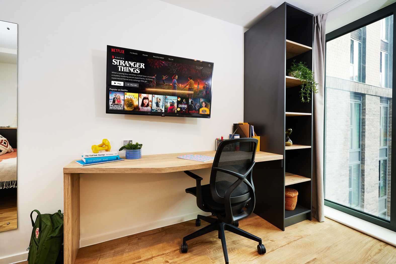 A modern study space with a wooden desk, black office chair, and a wall-mounted TV displaying "Netflix." There's a potted plant on a tall shelf, an open window revealing a city view, and books with a camera on the desk.