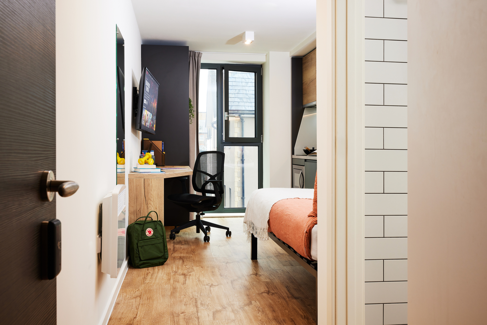 A modern dorm room with a bed, wooden floor, and a desk with a computer. A black chair is at the desk, and there's a green backpack on the floor. A window at the end of the room lets in natural light.