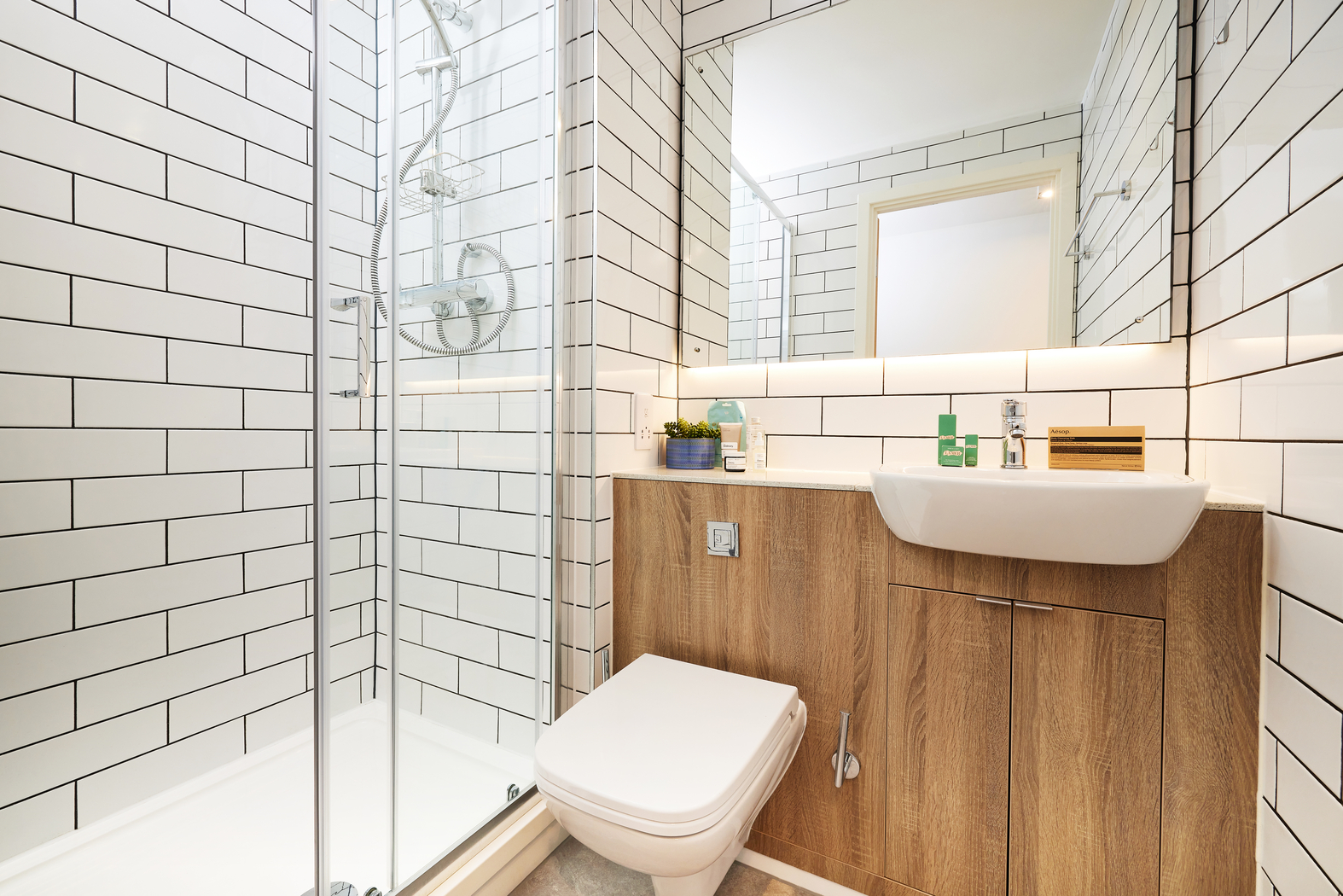Modern bathroom with a glass shower on the left, white subway tiles, a wooden cabinet below a sink with a rectangular mirror above, and a white toilet. A small plant and toiletries are on the countertop.