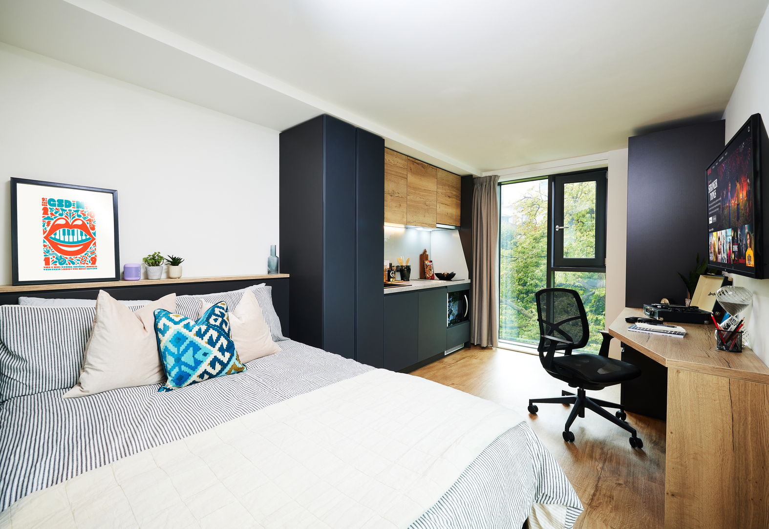 A modern bedroom with a bed featuring blue and white pillows and a light blanket. Dark cabinets and a small kitchenette are on one side. A desk with a chair and monitor sits near a window overlooking greenery. A colorful framed art piece is on display.
