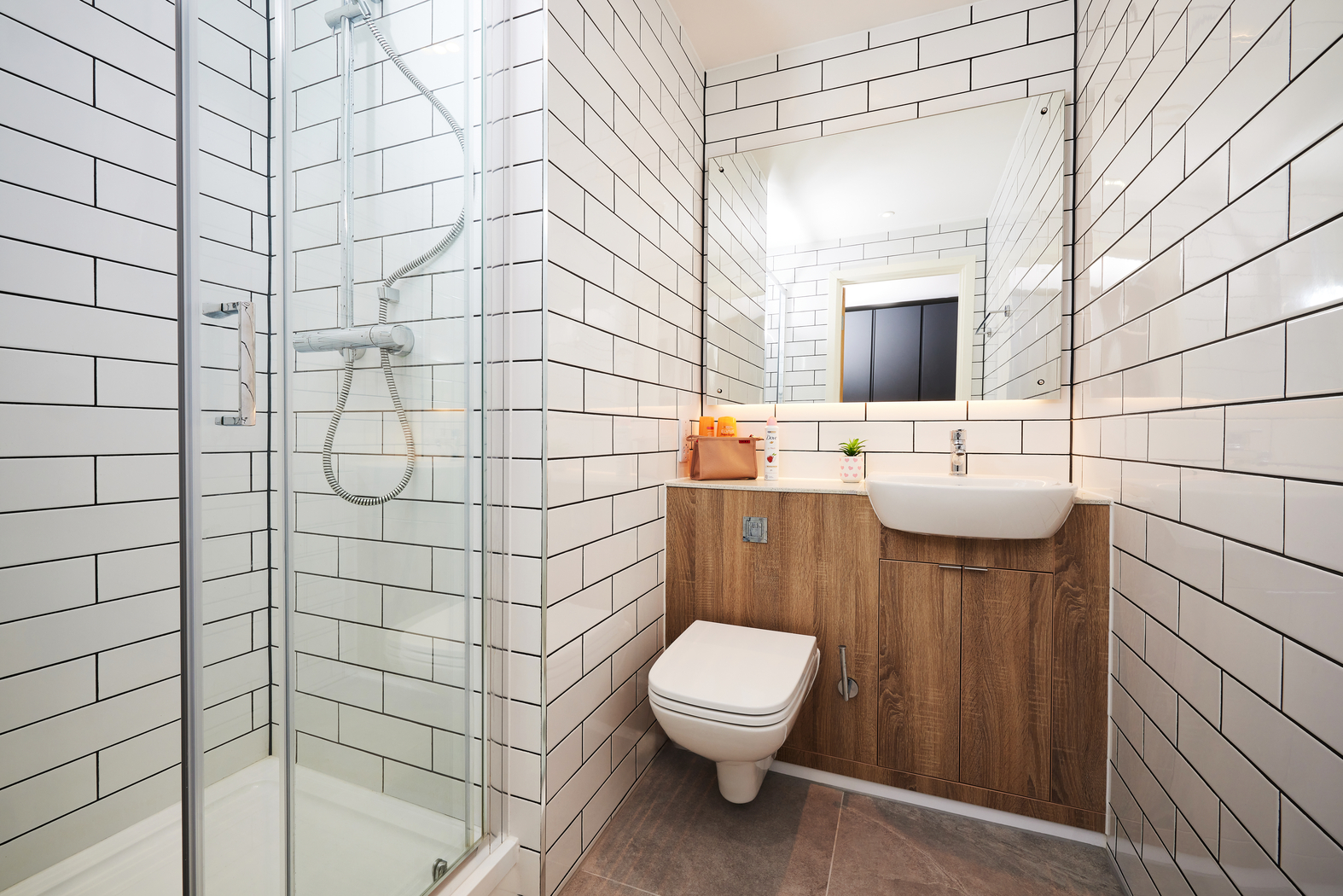 A modern bathroom with white subway-tiled walls and a glass-enclosed shower. There is a toilet and a wooden vanity with a white sink. A mirror reflects the door, and a small plant and toiletries are on the counter. The floor has grey tiles.