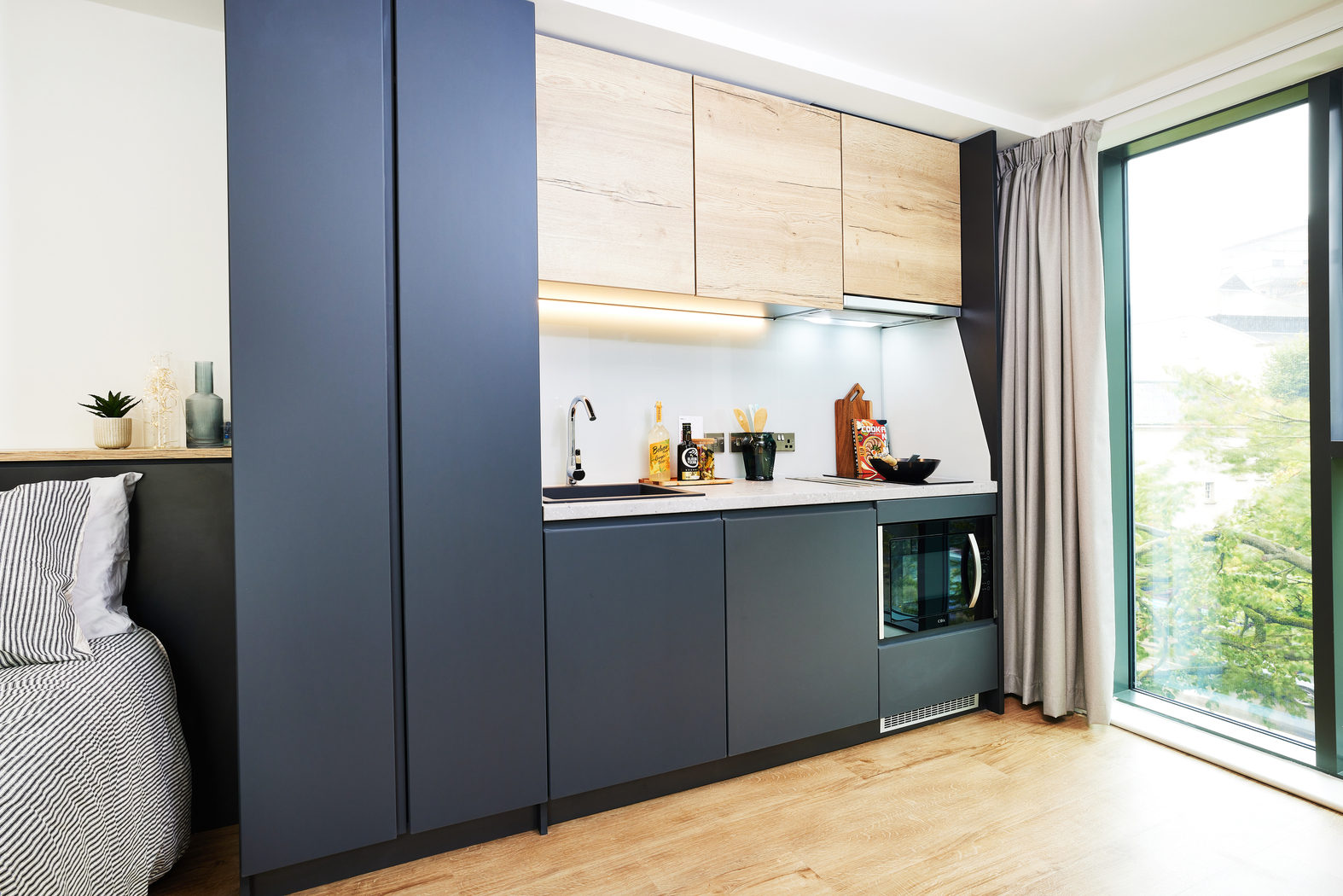 Modern studio apartment kitchenette with dark cabinets and wooden upper cupboards. Includes a sink, stove, and microwave. A large window with curtains on the right provides natural light. A bed with a striped cover is partially visible on the left.
