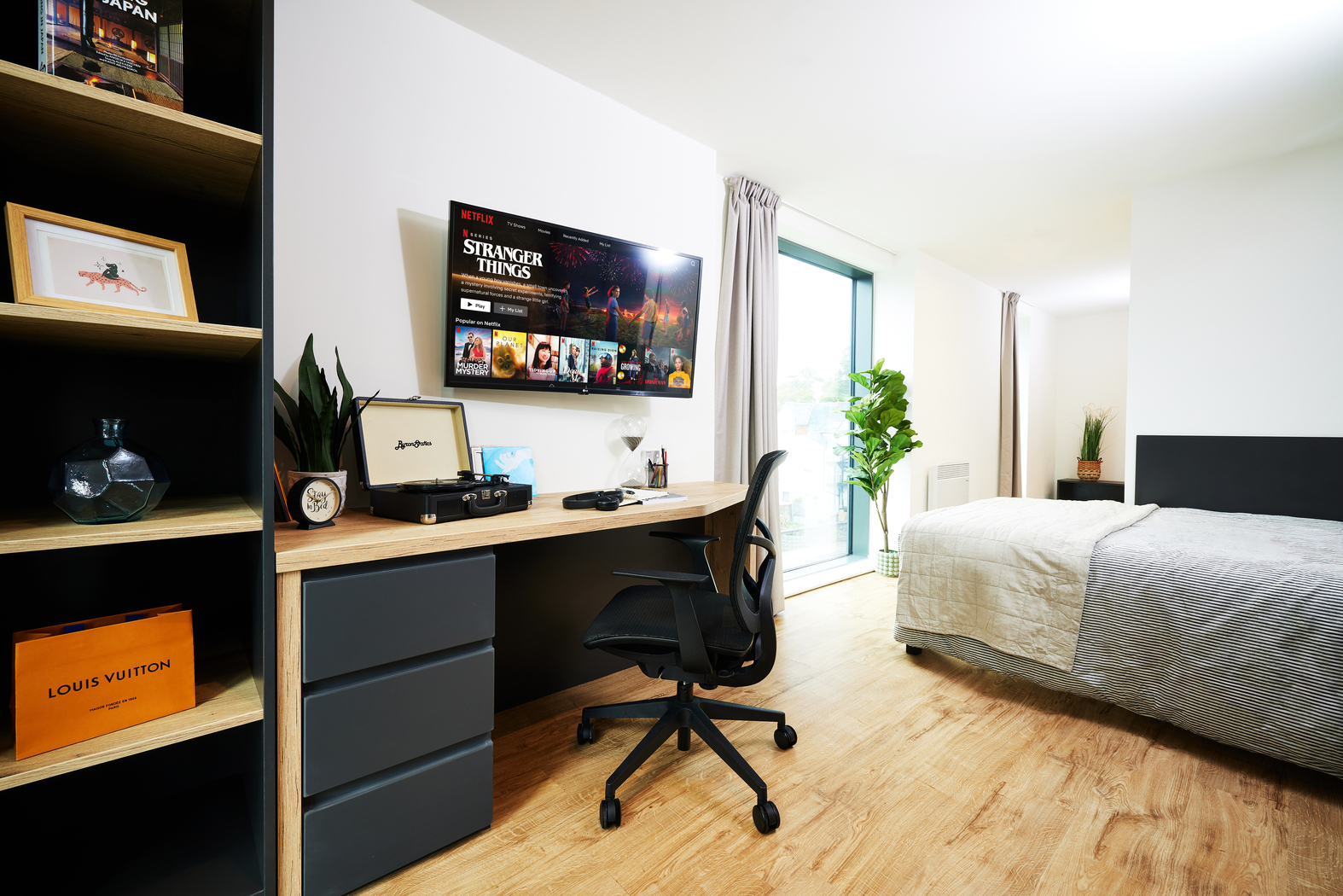 Modern bedroom with a stylish workspace. A desk holds a swivel chair, potted plants, framed art, and a TV screen displaying "Stranger Things." A bed with striped bedding is nearby. Spacious windows let in natural light, and shelves display decor items.