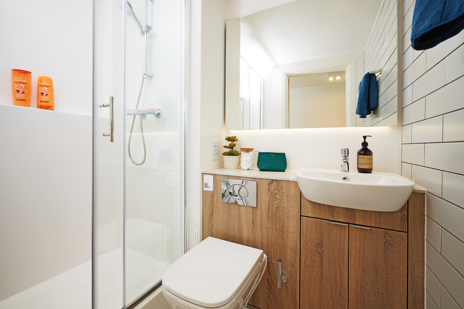 A modern bathroom with a glass-enclosed shower, wooden cabinet, and white sink. There are two orange bottles on a ledge in the shower. The wall has white tiles, and there's a mirror above the sink. A towel hangs on the right wall.