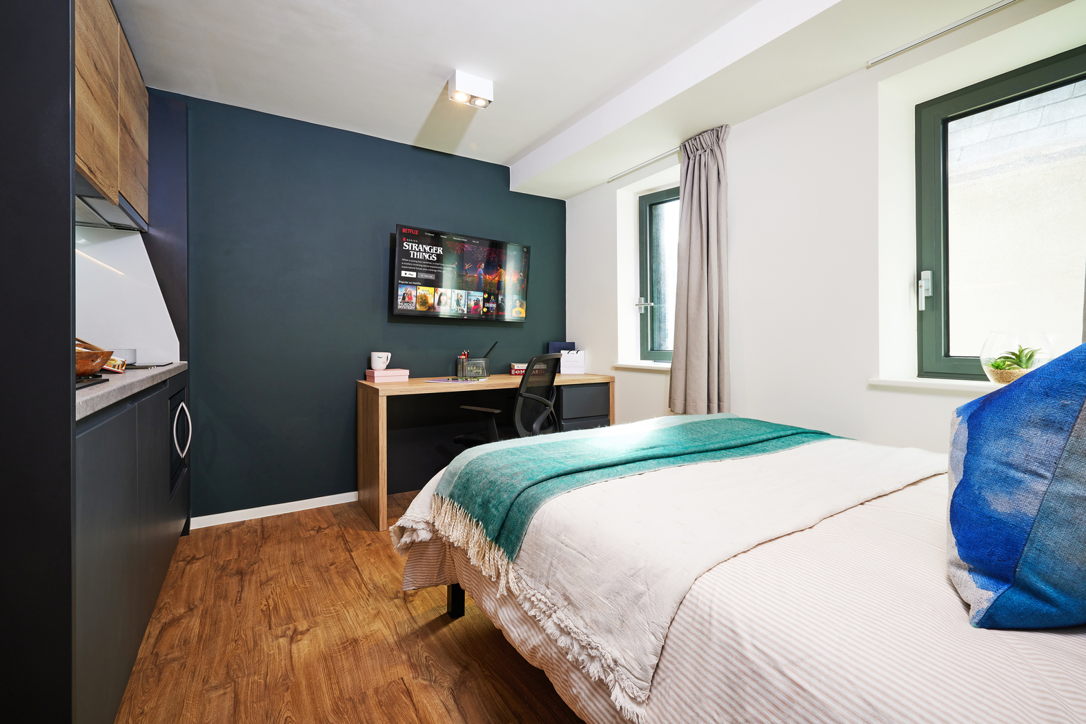 A cozy bedroom with a wooden floor features a neatly made bed with a white and teal blanket. A desk with a laptop and a television mounted on a dark accent wall are in view. Two windows with grey curtains let in natural light.