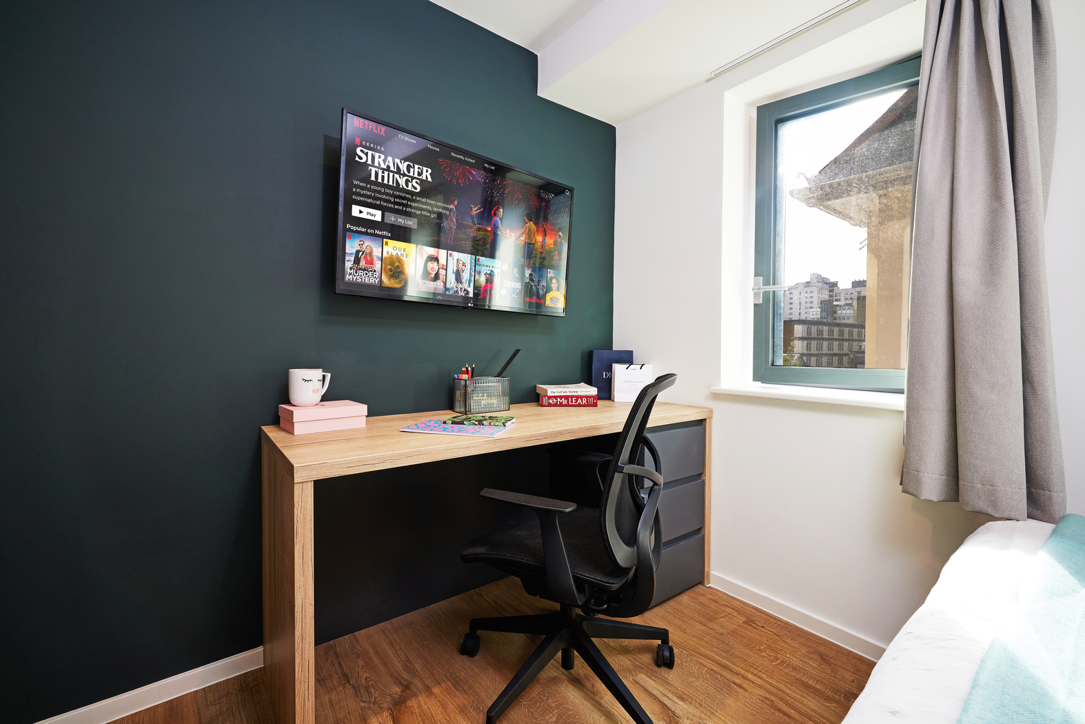 A modern study area in a bedroom with a black desk, a swivel chair, and a wall-mounted flatscreen TV displaying a streaming platform. The room features a window with light curtains and a hardwood floor. A bed is partially visible to the right.