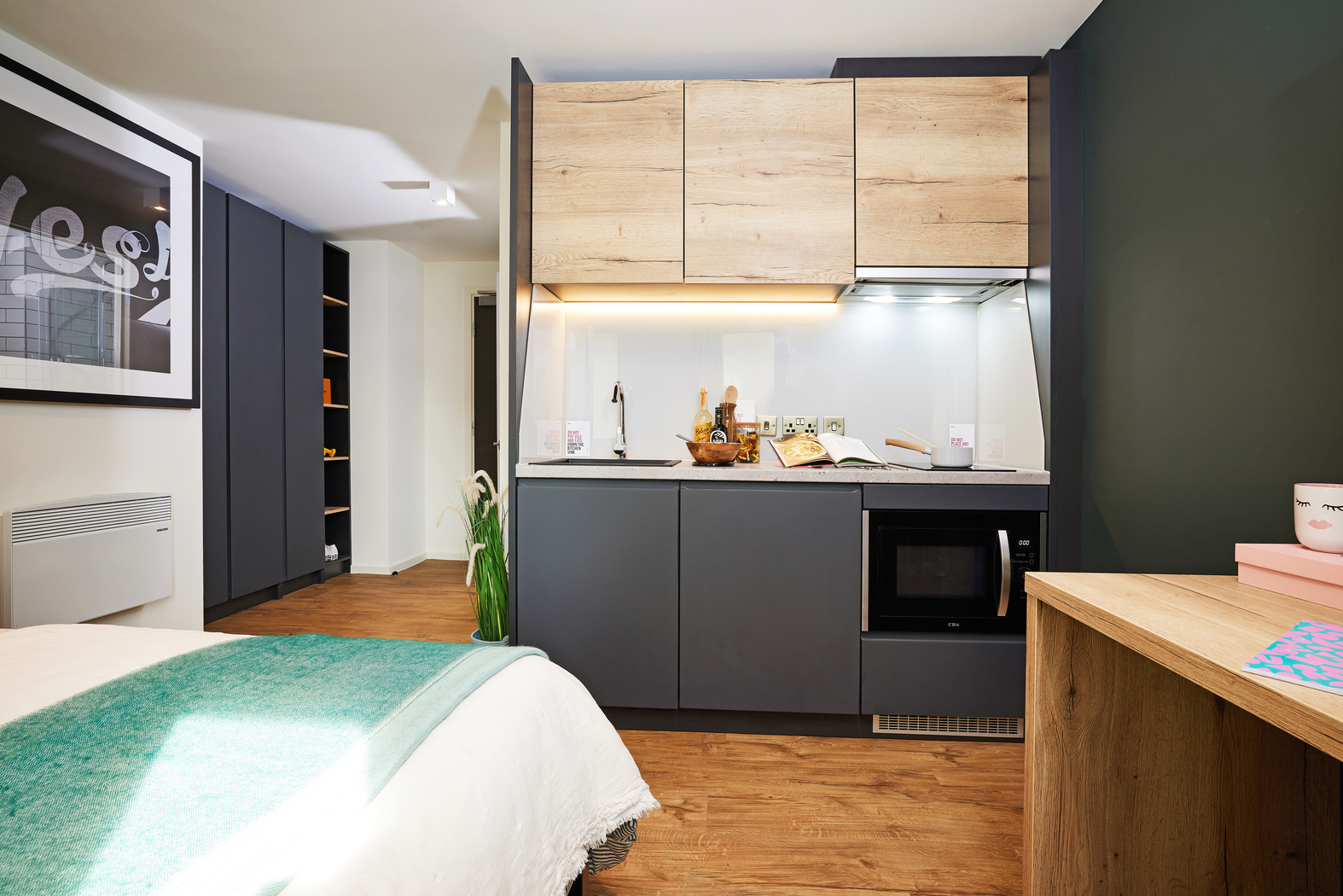 A modern studio apartment features a kitchenette with wood and gray cabinets. There's a microwave, a sink, and countertop items. A bed with a green throw, wall art, and a wooden desk are visible. The space has wooden flooring and a dark accent wall.