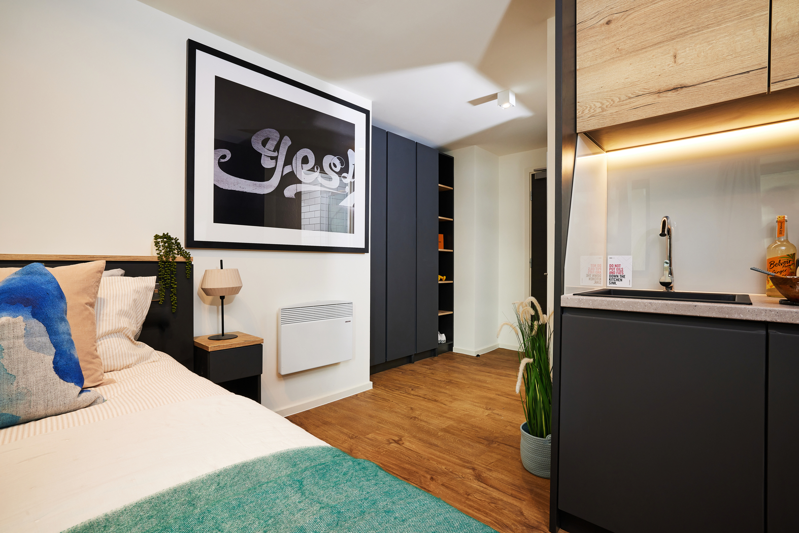 A modern studio apartment features a bed with blue and beige pillows, a large framed print that reads "Yes," a small wooden side table with a lamp, built-in cupboards, and a kitchenette with wood and black accents. A plant adds greenery to the space.