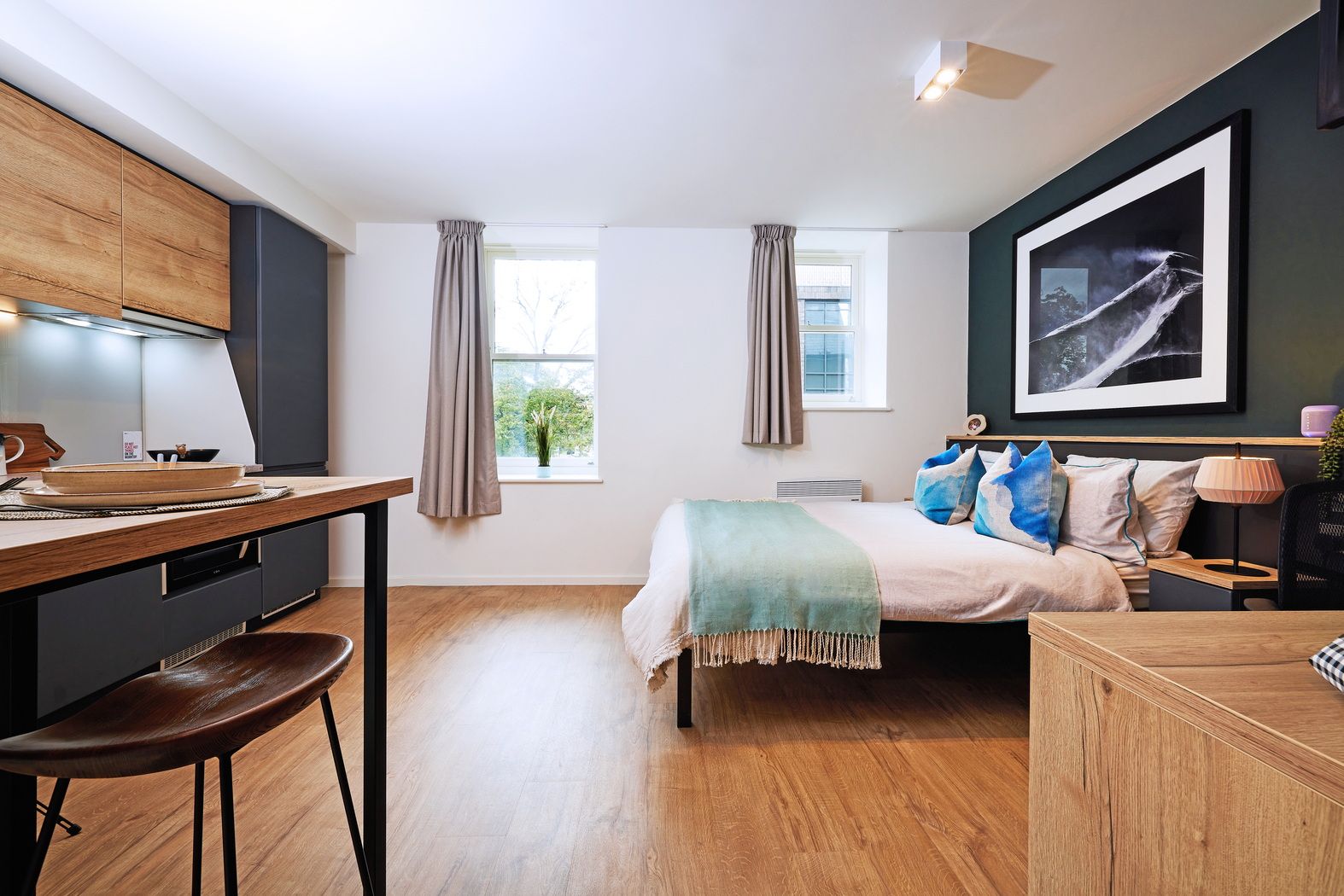 Modern studio apartment with a bed, pillows, and teal blanket. Two windows have gray curtains. A kitchenette features wooden cabinets, and a small dining table with a chair is nearby. A framed picture hangs on the dark wall above the bed. Wood flooring.