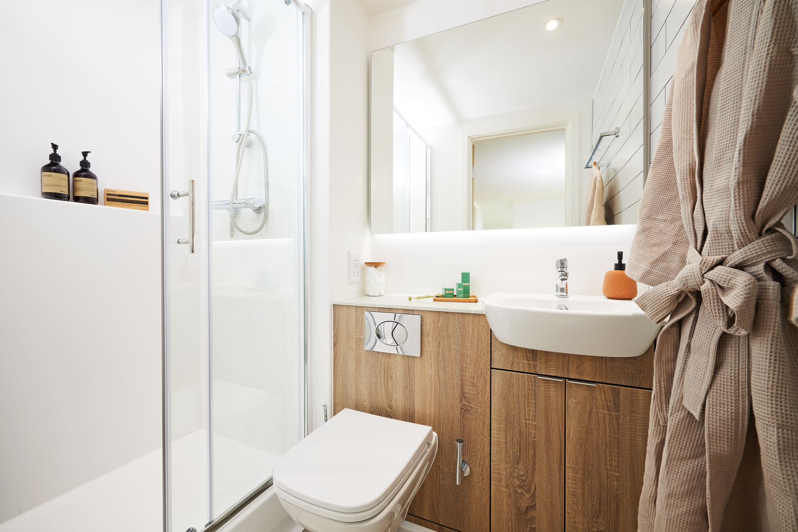 Small modern bathroom with wood-accented vanity, white sink, and wall-mounted toilet. Glass shower enclosure with chrome fixtures is on the left. Beige towels hang on hooks, and the mirror spans above the sink area.