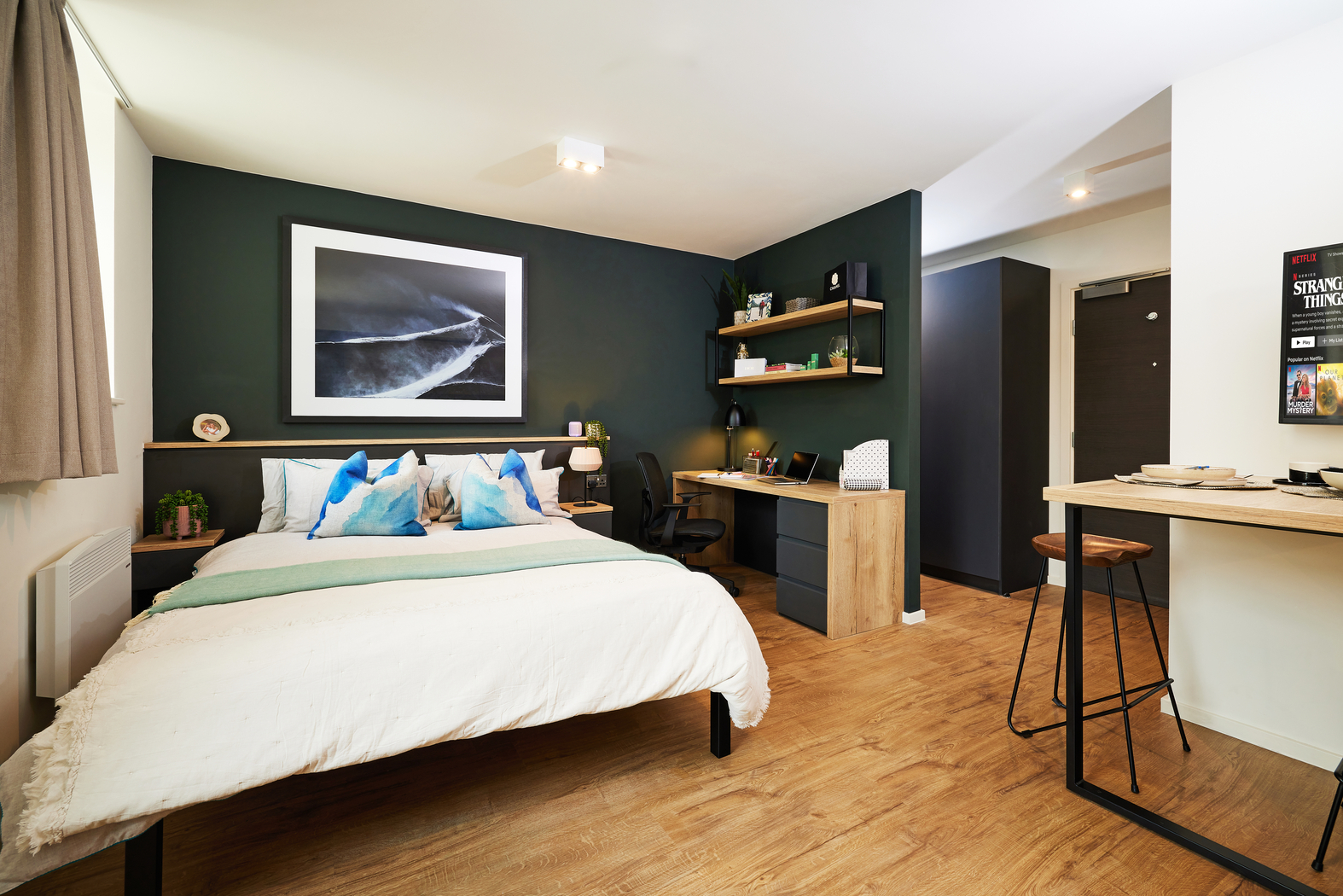 Modern bedroom with a cozy bed featuring blue and white pillows, a dark accent wall with a framed abstract photo, and a wooden desk with shelves. The room includes a wall-mounted TV and a breakfast bar with stools on a wooden floor.