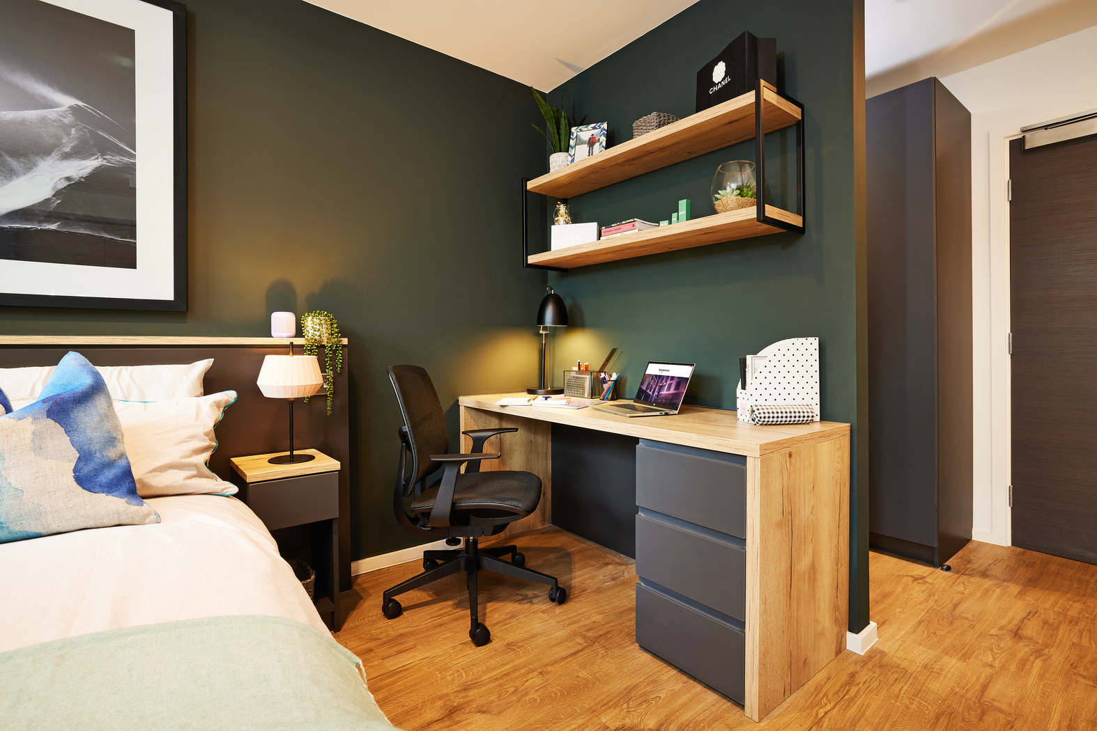A modern bedroom with a wooden desk and black chair. Shelves above the desk hold books and decor. A laptop and desk lamp sit on the desk. The bed with pillows is partially visible. Walls are dark green, and the floor is wooden.