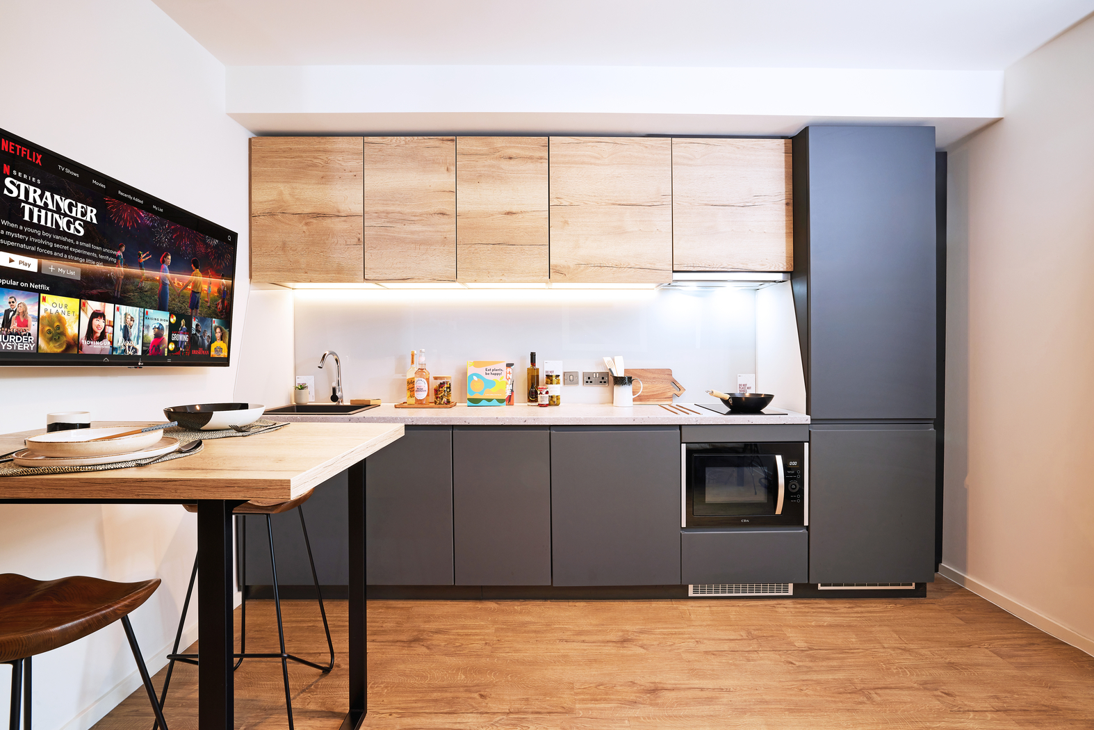 Modern kitchen with wood and gray cabinets, a wall-mounted TV showing streaming service content, a countertop with snacks and utensils, and a small dining table with a barstool. The floor is wooden, and the space is well-lit.