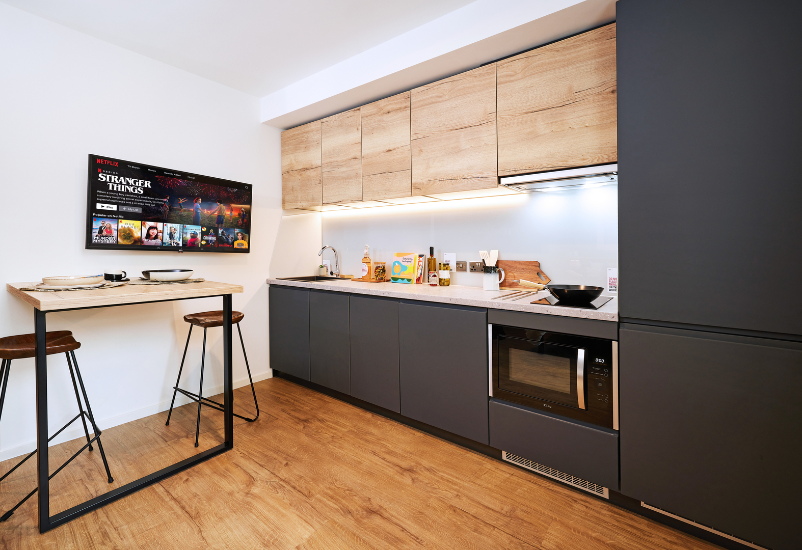 Modern kitchen with wooden flooring and cabinets, featuring a sleek counter with a microwave, cooking essentials, and a black stool table with plates. A wall-mounted TV is displaying a "Stranger Things" screen.