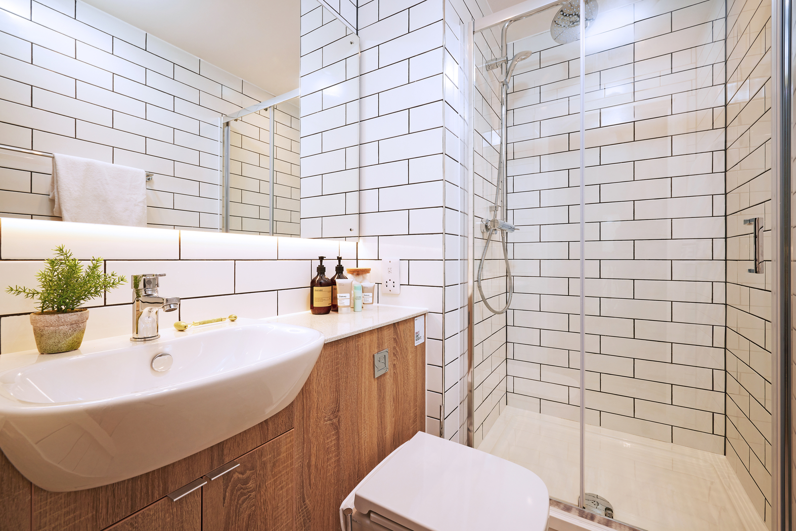 Modern bathroom with white subway tile walls, a glass-enclosed shower, a white sink on a wooden vanity, and a toilet. A small potted plant and toiletry bottles are on the countertop.