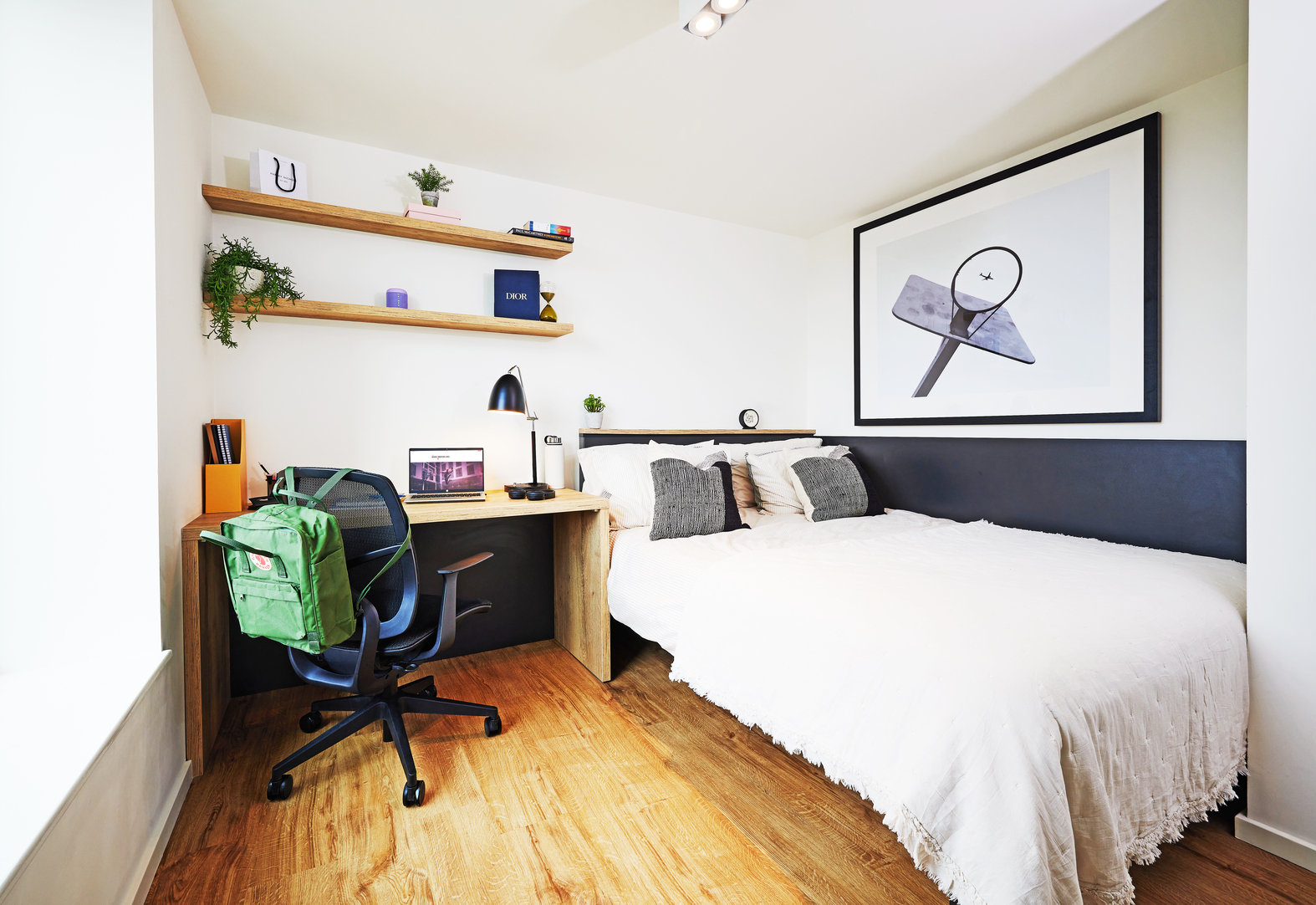 A cozy bedroom with a white bed, gray and black pillows, and a wooden desk. A laptop and lamp are on the desk with a green backpack on the chair. Shelves with decor and a large abstract art piece are on the walls. The floor is wooden.