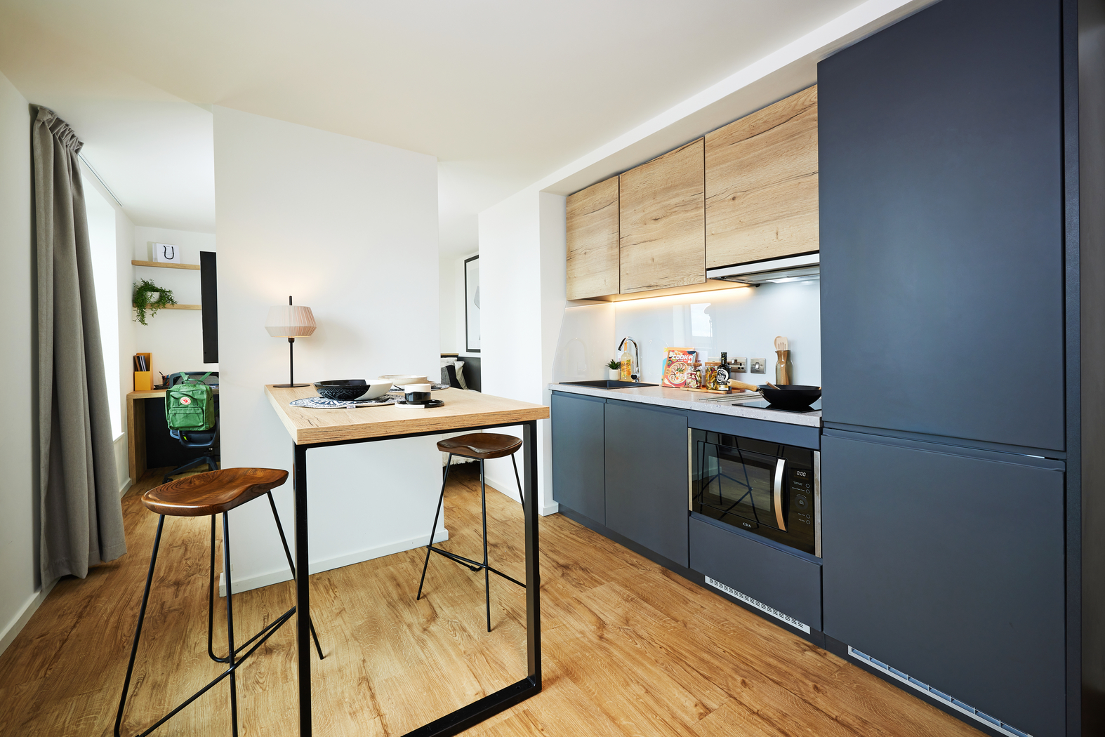 Modern kitchen with wooden flooring and cabinets, dark blue cabinetry, and a small dining area featuring a wooden table with metal legs and two high stools. A window with a curtain illuminates the room, and kitchen utensils are on the counter.