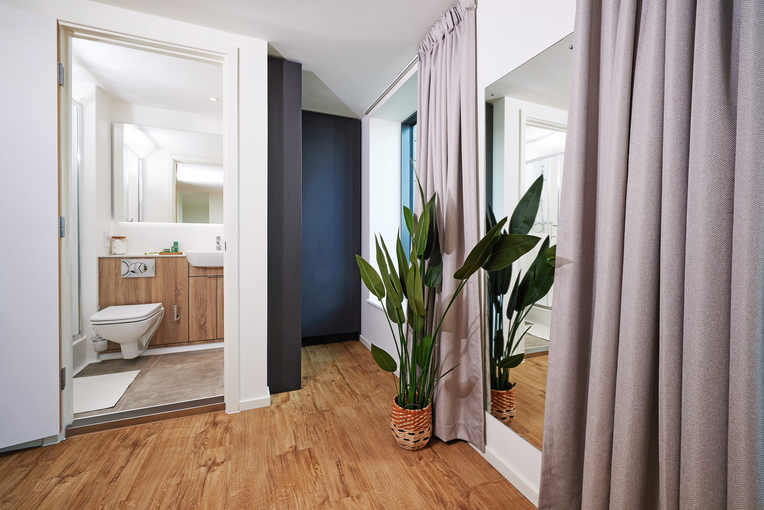 Modern bathroom interior with wood flooring leading to a bathroom. The bathroom features a wooden vanity and toilet. A large mirror and a potted plant with tall green leaves are placed on the left side of the entrance. Beige curtains are partially visible.