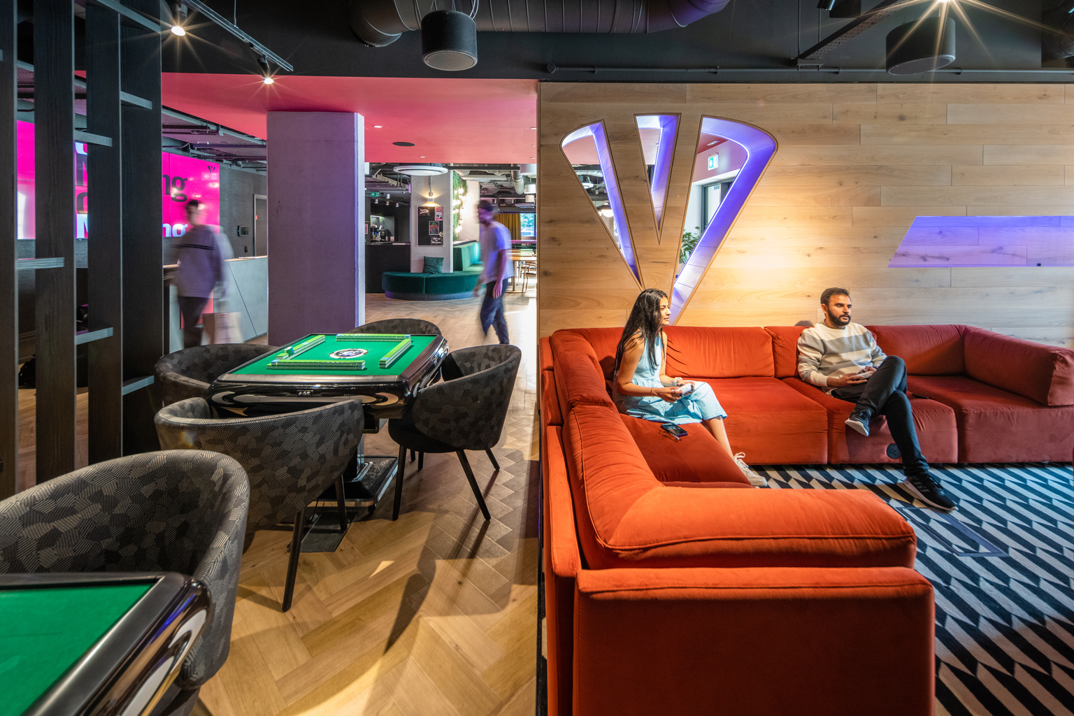 A modern lounge with a retro aesthetic features a red sectional sofa and two people conversing. Nearby, two gaming tables with green surfaces are surrounded by chairs. Neon lights and wood paneling add vibrant touches to the open space.