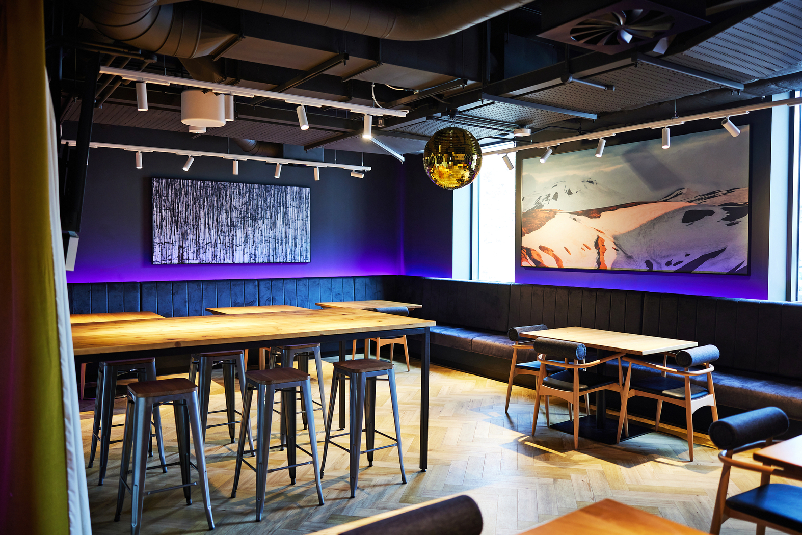 A modern dining space with wooden tables and stools. The room features a wooden floor, abstract paintings, purple accent lighting, and a reflective disco ball hanging from the ceiling. Large windows let in natural light.