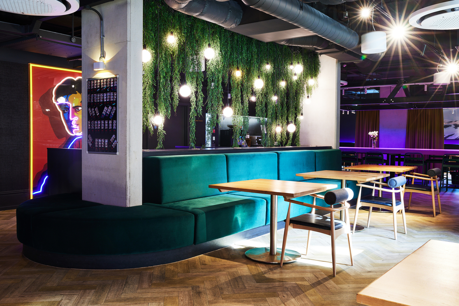 Chic modern lounge with teal velvet booths, wooden tables, and chairs. Ceiling lights hang over a wall lined with leafy greenery. A neon art piece of a face glows in the dim background. The setting exudes a trendy, cozy atmosphere.