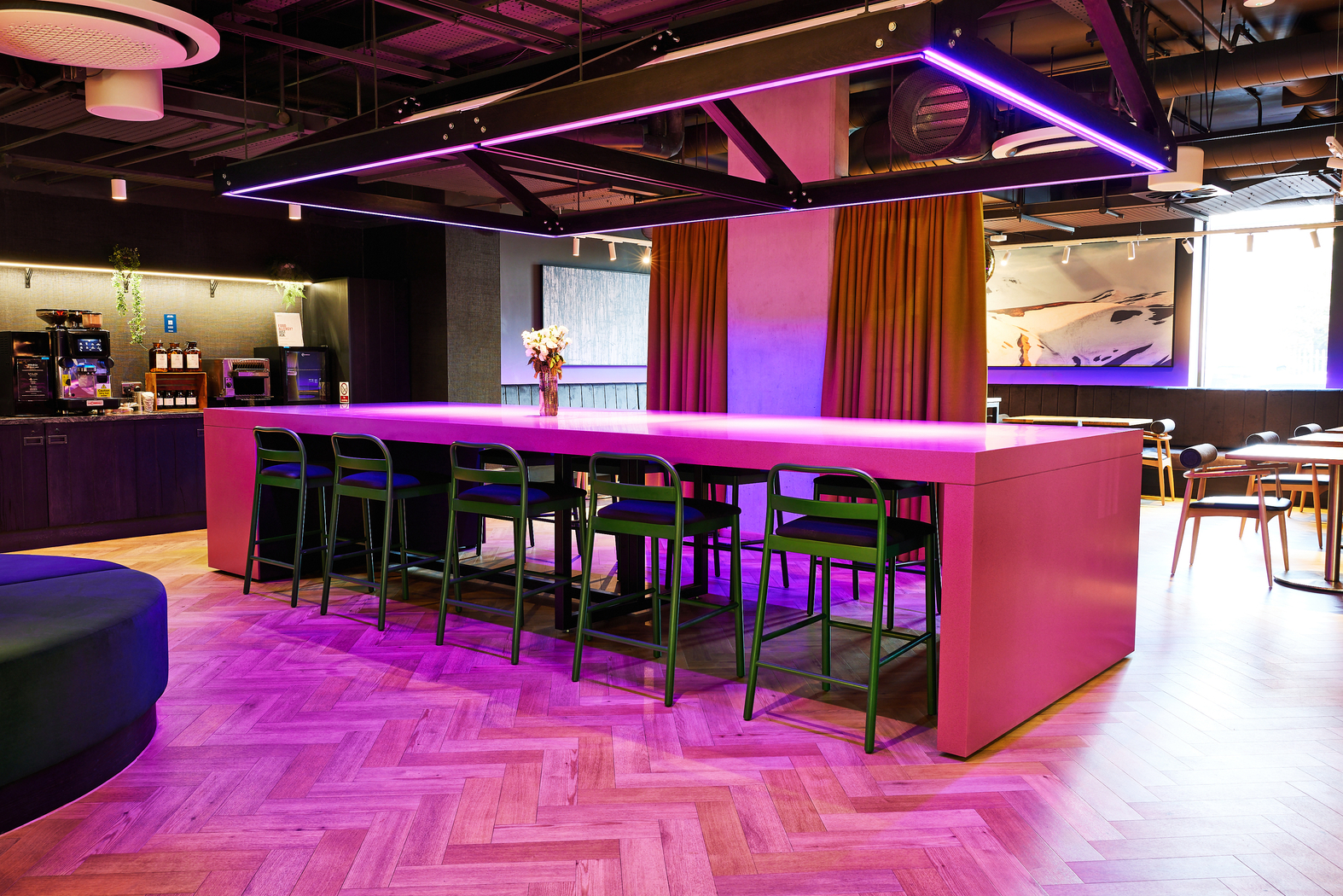 A modern lounge with a long pink table surrounded by black chairs under purple neon lighting. The setting includes a wooden floor, colorful curtains, a coffee station, and artwork on the wall, creating a trendy and inviting atmosphere.
