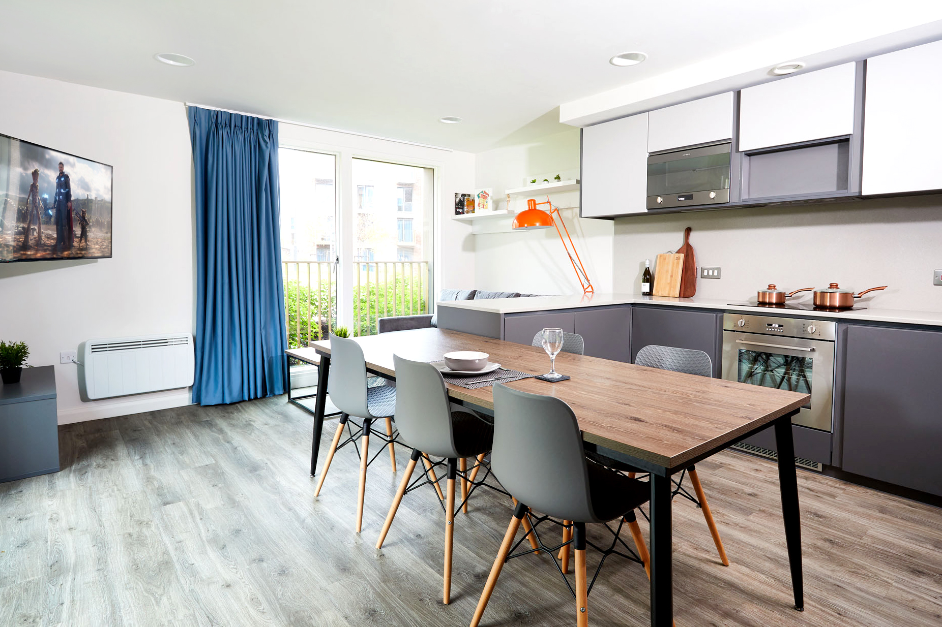 A modern kitchen-dining area on Lawrence Street features gray cabinets, a wooden dining table, and four gray chairs. Large windows with blue curtains flood in natural light. A wall-mounted TV is on the left, and orange and white accents adorn the kitchen counter.