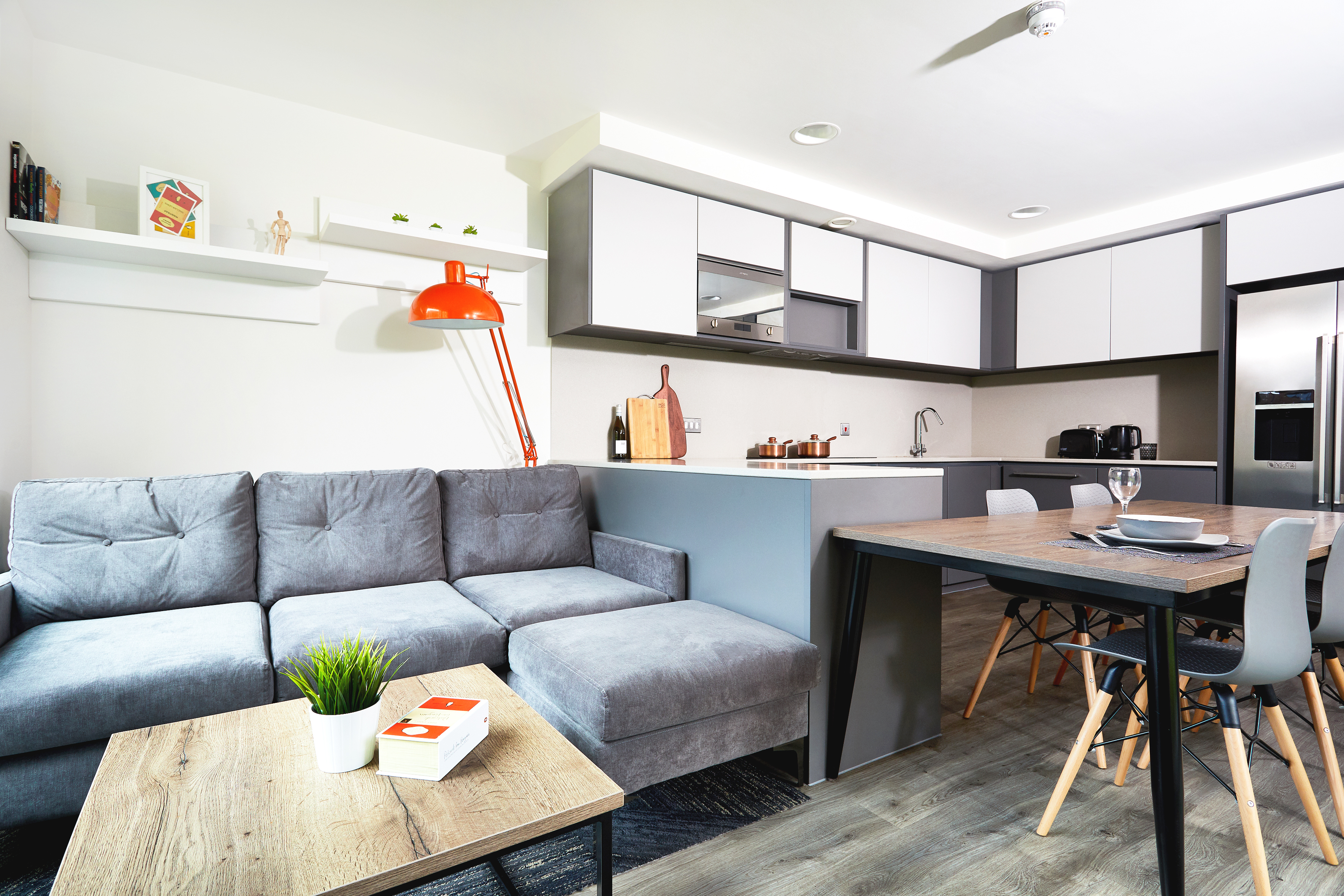 Modern open-plan living space on Lawrence Street features a gray sectional sofa, wooden coffee table, and vibrant orange floor lamp on the left. On the right, discover a kitchen with gray and white cabinetry and a dining area equipped with a wooden table and gray chairs.