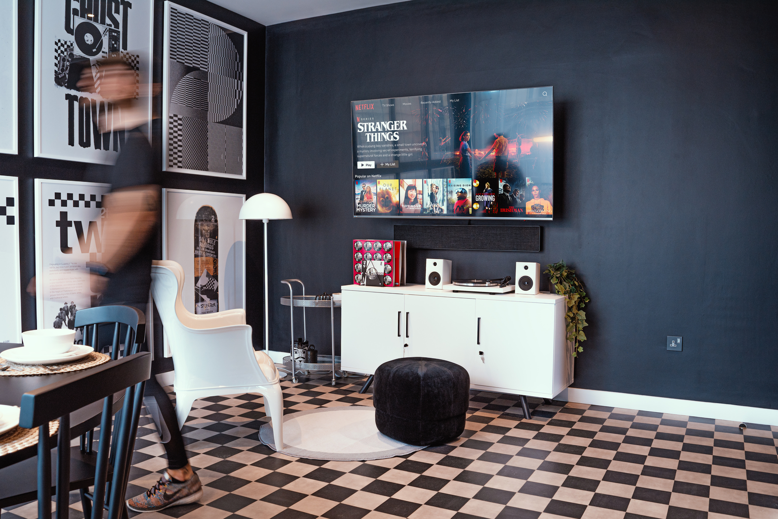 In a modern living room with black-and-white decor, a wall-mounted TV displays Netflix. A person walks by, blurred in motion. A white chair, round ottoman, and cabinet with speakers accent the checkered floor, while framed art highlights architecture themes and Copper Towers.