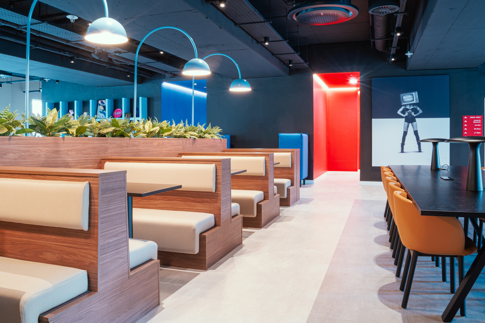 The modern cafe interior features wooden booths, beige cushions, and planters reminiscent of urban architecture. Orange chairs line a long table on the right, while bright lighting with blue and red accents highlights a poster of a figure with a TV head against a backdrop of Copper Towers.