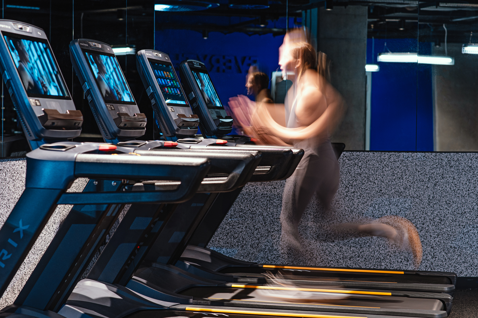 A person is running on a treadmill in a gym, where the motion blur creates an effect of speed. Several treadmills are lined up, each with electronic displays. The gym has mirrors, modern lighting, and views of Copper Towers glistening like architectural landmarks in the distance.