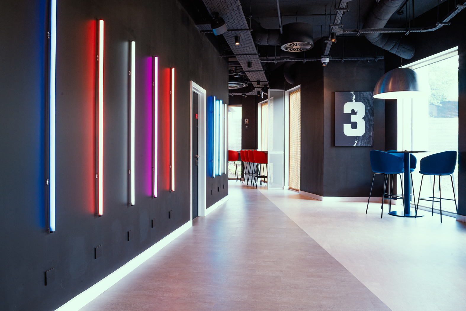 A modern hallway with glowing neon lights on a dark wall showcases stunning architecture. Bar stools and a high table sit near large windows, while a black and white artwork featuring the number 3 hangs in the background. Ceiling pipes and track lights add an urban touch to this stylish space.