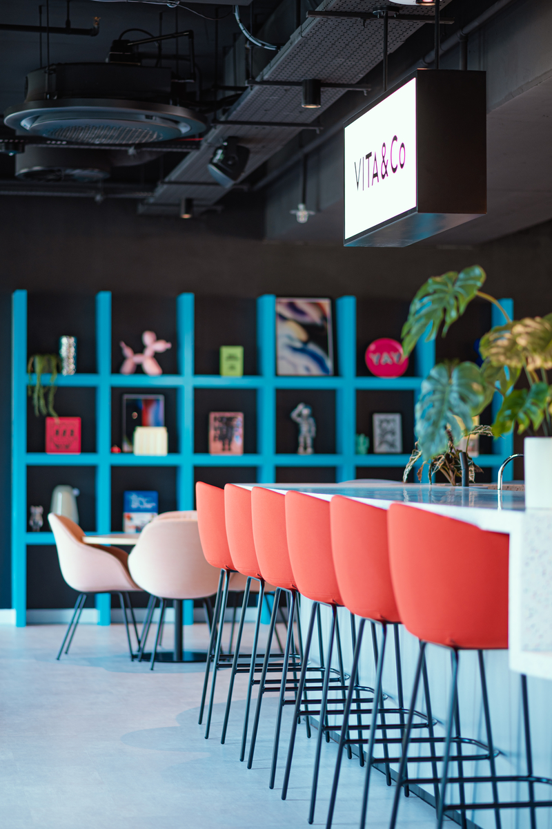 Modern office lounge in a stylish student accommodation in Coventry, featuring a row of red bar stools at the counter. Behind, there's a shelving unit with colorful decor. A small table with white chairs sits on the left. Ceiling lights and a hanging sign reading "VITA & Co" illuminate above.