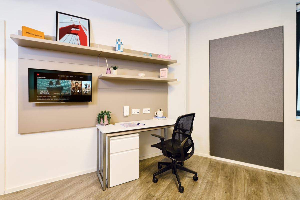 A modern, minimal home office with a desk, black office chair, wall-mounted TV, floating shelves with decor, and a large pinboard on the wall. The space is tidy and well-lit with wooden flooring.