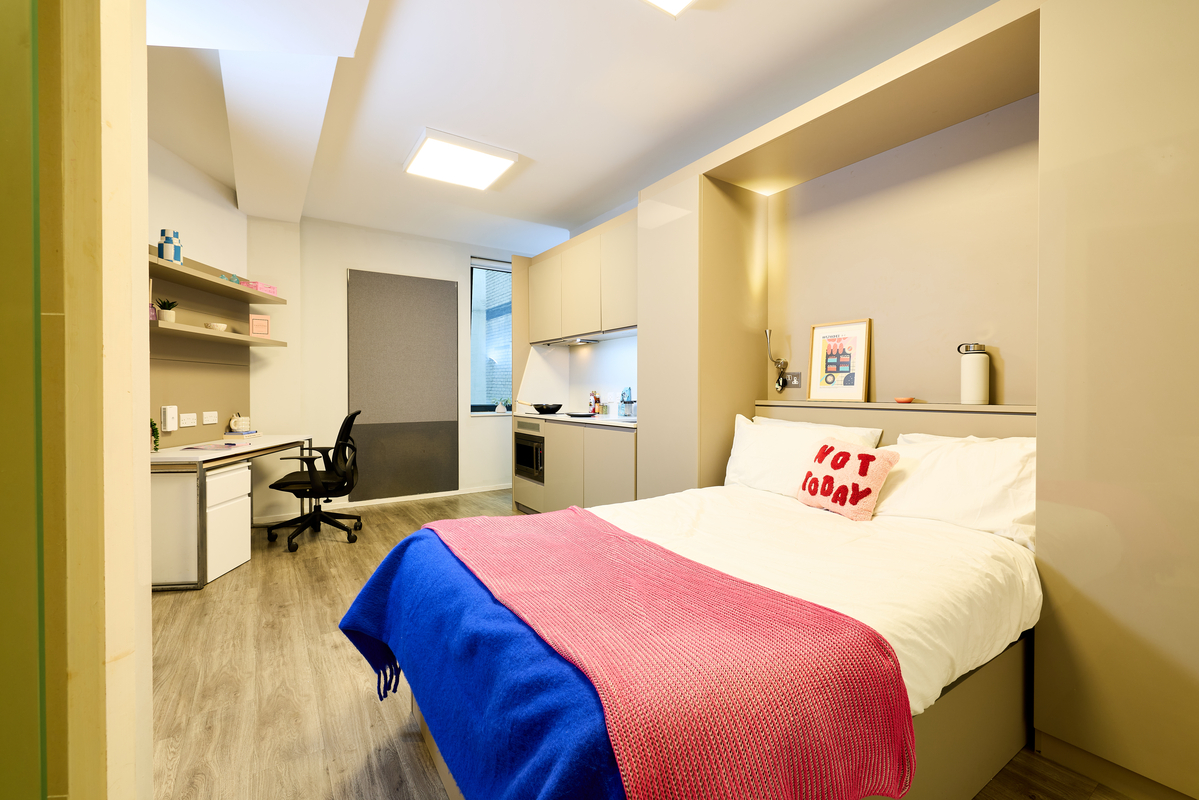 A modern studio apartment with a neatly made bed, colorful blankets, a desk with a chair, open shelves, a small kitchenette, and large window, all set on light wood flooring.