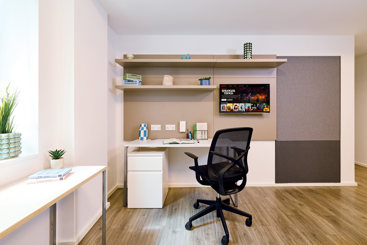 A modern home office with a white desk, black mesh chair, wall-mounted shelves, a small TV, potted plants, and wooden flooring. Natural light enters through a window on the left.