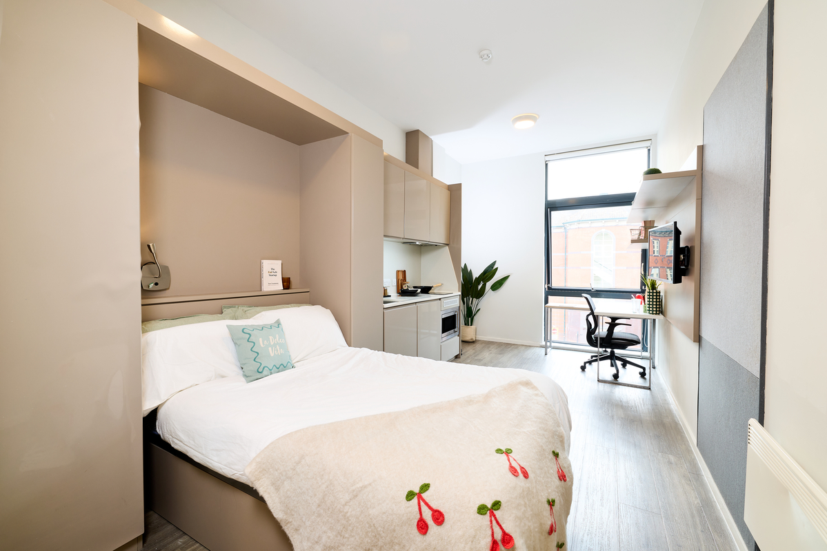 A modern, bright studio apartment in Portland House with a bed, kitchenette, desk, chair, wall-mounted TV, and large window. Minimal decor features light colors, a plant by the window, and a cherry-patterned blanket on the bed.