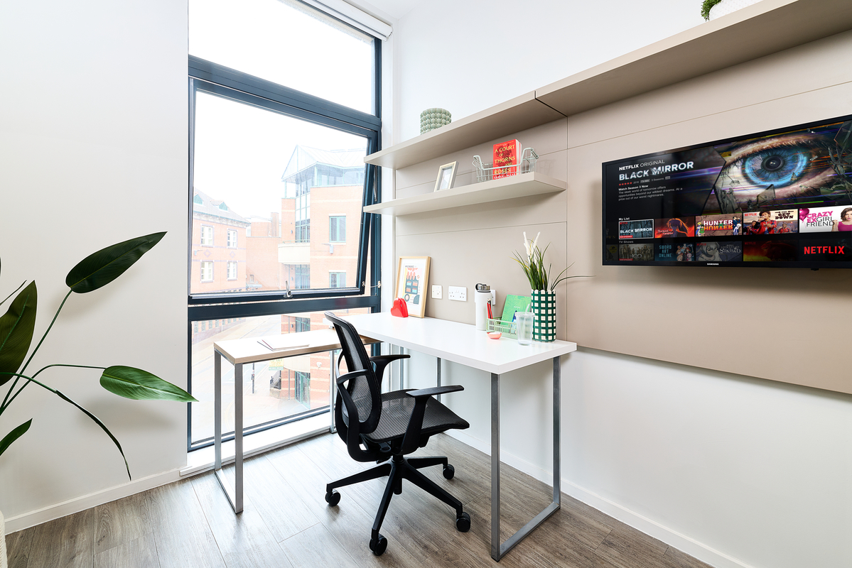 Modern, bright study room in Portland House with a desk, office chair, and shelves. A large window lets in natural light; a TV on the wall displays Netflix. Decor includes plants, books, and minimalist accents.