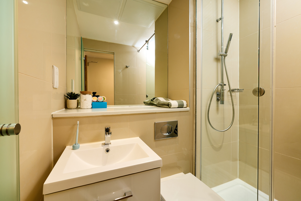 Modern bathroom at Portland House with beige tiles, a glass shower enclosure, a white sink with a blue toothbrush, and a shelf holding a plant, toiletries, and neatly folded towels—all reflected in a large mirror.